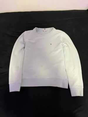 Ljusblå sweatshirt från Tommy Hilfiger - Ljusblå sweatshirt med rund hals och långa ärmar från Tommy Hilfiger. Tröjan har en liten broderad logga på bröstet och ribbade muddar vid ärmslut och nederkant. Perfekt för en avslappnad och stilren look.