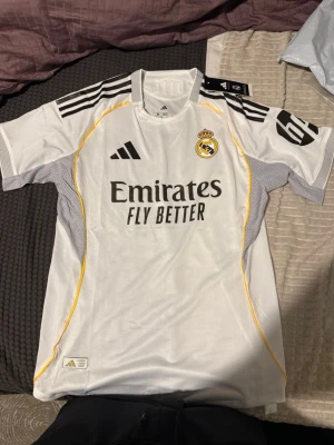 Real Madrid matchtröja Adidas vit - Säljer en vit Real Madrid matchtröja från Adidas med svarta detaljer och gula linjer längs sidorna. Tröjan har klubbmärke på bröstet, Adidas-logga och sponsortryck 'Emirates Fly Better' framtill. Tillverkad i lätt och ventilerande material, perfekt för fotboll.