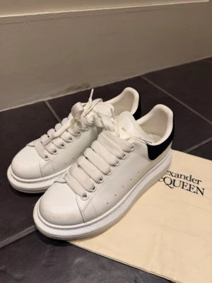 Sneakers inför hösten🌸 - Stilrena vita sneakers från Alexander McQueen med svart mockadetalj på hälen och guldfärgad logga. Skorna har tjock sula, breda vita skosnören och perforerade sidor för ventilation. Perfekta för dig som gillar en clean och modern look.