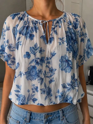 Blommig blåvit blus från My Wear - Söt blus i tunn, luftig kvalitet med blått blommönster på vit botten. Modellen har korta ärmar, rund halsringning med knytband och lätt rynkad detalj vid halsen. Perfekt till jeans för en fräsch och somrig look.