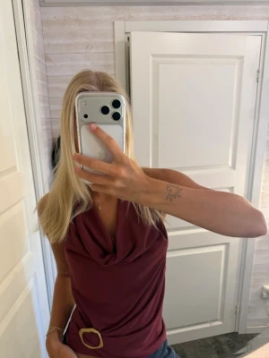 Vinröd ärmlös topp med öppen rygg - Snygg vinröd topp med draperad halsringning och öppen rygg. Toppen är ärmlös och har ett dekorativt bälte i midjan. Perfekt för dig som vill ha en trendig och stilren look.