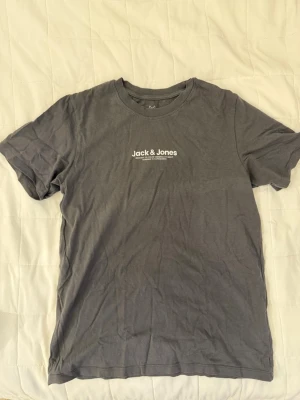 Grå t-shirt från Jack & Jones - Säljer en enkel och stilren grå t-shirt från Jack & Jones med vit logga tryckt på bröstet. T-shirten har rund halsringning och korta ärmar, perfekt till vardagslooken. Materialet känns mjukt och skönt mot huden.