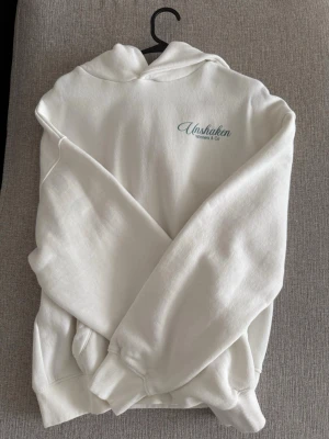 Vit hoodie med tryck från Bershka  - Säljer en vit hoodie från Unshaken med huva och ribbade muddar. På bröstet står det 'Unshaken Winners & Co' och på ryggen finns ett stort tryck med texten 'WIN WHEN YOU CHILL' i rosa och blått samt tärningsgrafik. Perfekt för en avslappnad stil.