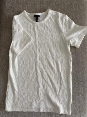 Vit strukturerad t-shirt med mönster - Snygg vit t-shirt med diskret strukturmönster och rund hals. T-shirten har korta ärmar och är gjord i ett mjukt, stretchigt material som ger en bekväm passform. Perfekt för dig som gillar stilrena och enkla plagg med en twist.