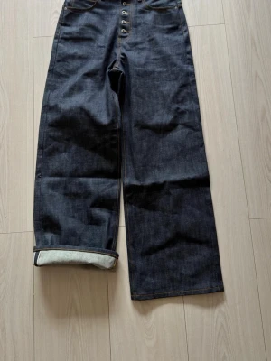 No faith studios selveged denim raw denim bootcut jeans - Säljer ett par mörkblå selvedge jeans med hög midja och raka, vida ben. Jeansen har knäppning framtill och en uppvikt kant på ena benet. Tillverkade i 100% bomull för en klassisk denimkänsla.