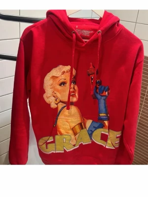 Röd HAVOC hoodie med Grace-tryck - Snygg röd hoodie från HAVOC med stor färgglad Grace-illustration på framsidan. Hoodien har huva med dragsko och tryck i gult, blått och beige. Perfekt för dig som vill sticka ut med en unik och kreativ look.