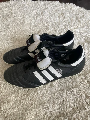 Adidas Copa Mundial fotbollsskor svart/vit - Använt vid endast 3 tillfällen, som nya! Nypris 1300kr. Klassiska Adidas Copa Mundial fotbollsskor i svart läder med vita detaljer och de ikoniska tre ränderna på sidan. Skorna har svart snörning, vit plös med Adidas-logga och låg profil. Perfekta för gräsplaner och dig som vill ha tidlös stil på planen. 