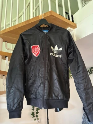 Svart bomberjacka från Adidas - Snygg svart bomberjacka från Adidas med vit logga och tryck på bröstet samt röd patch. På ryggen står det 'New York's Finest' i stora bokstäver. Jackan har ribbade muddar och dragkedja framtill. Perfekt för dig som gillar streetwear och sportig stil.