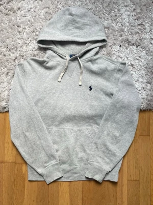 Grå hoodie från Polo Ralph Lauren - Ljusgrå hoodie från Polo Ralph Lauren med klassisk marinblå logga broderad på bröstet. Hoodien har dragsko i huvan, magficka och ribbade muddar vid ärmar och nederkant. Tillverkad i mjuk bomullsblandning för extra komfort.