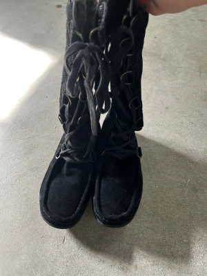 Mockastövlar med snörning - Säljer ett par svarta stövlar i mocka med snörning framtill och dragkedja på sidan. Stövlarna har rund tå och mjukt foder på insidan, vilket gör dem perfekta för kyligare dagar. Snygg design med dekorativa metallringar längs snörningen.Strl 36, används endast en gång.