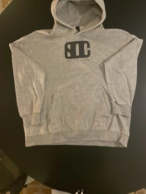 Grå hoodie från SOC med tryck - Säljer en grå hoodie från SOC med svart logga och texten 'SPORT OF CLASS' på bröstet. Hoodien har huva, magficka och ribbade muddar vid ärmar och nederkant. Perfekt för en avslappnad och sportig stil.