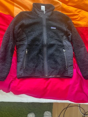 Svart teddyjacka från Patagonia - Mysig svart teddyjacka från Patagonia med dragkedja framtill och två fickor med dragkedja. Jackan har hög krage och paneler i slätare material på sidorna och ärmarna för extra stil och komfort. Perfekt för kyliga dagar och enkel att matcha.