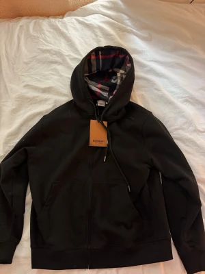 Svart hoodie med Burberry-mönstrad huva - Snygg svart hoodie med dragkedja från Burberry. Huvan har det klassiska Burberry-rutiga fodret i beige, rött och svart. Hoodien har känguruficka framtill och justerbara snören i huvan. Perfekt för en stilren och avslappnad look.