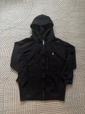Svart Ralph lauren hoodie  - Säljer en svart hoodie med dragkedja och huva. Hoodien har snören i huvan, två fickor framtill och en liten vit logga broderad på bröstet. Perfekt för en chill och avslappnad stil. Materialet känns mjukt och bekvämt.