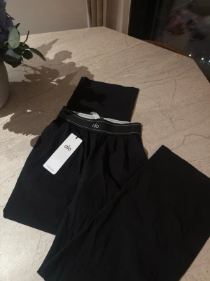 Svarta byxor från Alo, storlek S - Snygga svarta byxor från Alo med hög midja och elastisk linning. Tillverkade i ett mjukt och stretchigt material som sitter skönt på kroppen. Perfekta för träning eller chill. Diskret Alo-logga på linningen och stilren design. Oanvända 