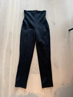 Svarta högmidjade byxor - Snygga svarta byxor med hög midja och raka ben. Byxorna har en stilren look med markerade sömmar framtill och dragkedja i ryggen. Perfekta för dig som gillar en clean och modern stil.