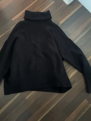 Svart polotröja i stickat material - Mysig svart stickad polotröja med bred ribbad kant nedtill och vid halsen. Tröjan har en avslappnad passform och långa ärmar, perfekt för kyliga dagar. 