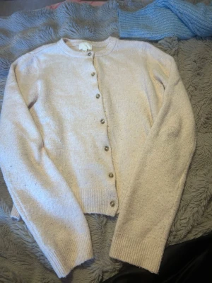 Ljusrosa kofta med pärlemorknappar - Mysig ljusbeige kofta med rund halsringning och pärlemorskimrande knappar framtill. Koftan har långa ärmar och ribbade muddar vid ärmslut och nederkant. Perfekt att slänga över en topp för extra värme.