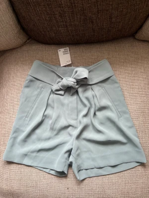 Ljus turkos  shorts med knyt från H&M - Snygga ljus turkos shorts från H&M med hög midja och brett knytband i midjan. Shortsen har två sidofickor och en lös passform som ger en avslappnad look. Perfekta för varma dagar och enkla att matcha med olika toppar.