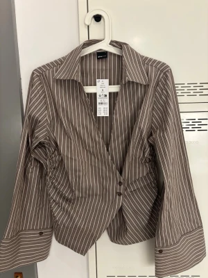 Slutsåld wrap shirt från Gina Tricot - Säljer en beige och vit randig skjorta från Gina Tricot i omlottmodell. Skjortan har långa ärmar med knappdetaljer vid manschetterna och klassisk krage. Slutsåld på Gina Tricots hemsida, säljer då jag ej hunnit returnera i tid 🩷