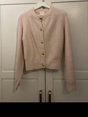 Ljusrosa ribbad kofta med guldknappar - Supersöt ljusrosa kofta med ribbad struktur och guldiga knappar framtill. Kortare modell med rund halsringning och långa ärmar. Perfekt att styla över en topp eller klänning för en trendig look.