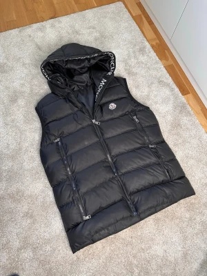 Svart Moncler väst  - Snygg svart dunväst från Moncler med avtagbar huva och dragkedja framtill. Storleken är L och passar dig från 175-185. Västen har två praktiska fickor med dragkedja och Moncler-logga på bröstet. Perfekt för lager-på-lager och håller dig varm med sin vadderade design. Skriv vid intresse, pris kan diskuteras🔥🔥🔥