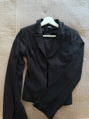 Svart kavaj Gina Tricot XS - Stilren svart kavaj från Gina Tricot i storlek XS. Klassisk modell med smal passform, spetsiga slag och knäppning framtill. Perfekt att matcha med skjorta eller t-shirt för en snygg look. Materialet känns mjukt och följsamt. Slits vid ärmarna. 