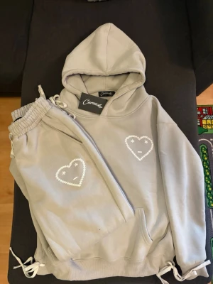 Grått loungewear-set från Carsicko - Mysigt grått loungewear-set från Carsicko med hoodie och matchande byxor. Båda plaggen har ett vitt hjärta med broderad smiley på bröstet och benet. Setet har dragsko i midjan och nederkant samt mjukt material som passar perfekt för chill dagar. PRIS KAN DISKUTERAS.