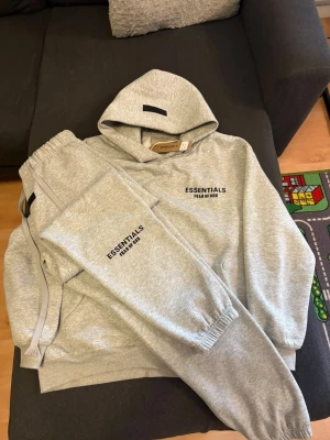 Grått loungewear-set Essentials S - Säljer ett grått loungewear-set från Essentials Fear of God i storlek S. Setet består av en hoodie med huva och matchande byxor med resår i midja och benslut. Båda plaggen har svart Essentials Fear of God-tryck på bröst och ben. Setet kommer även med original taggen för både byxorna och huvtröjan. Perfekt för dig som vill sticka ut med din stil. PRIS KAN DISKUTERAS.