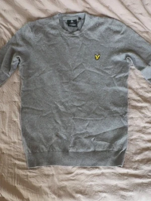 Grå stickad tröja från Lyle & Scott - Snygg grå stickad tröja från Lyle & Scott med rund halsringning och den klassiska gula loggan broderad på bröstet. Tröjan har ribbade muddar vid ärmslut och nederkant. Perfekt till lager på lager och enkel att matcha.