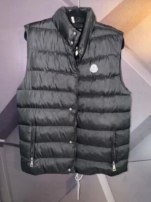 Moncler väst - Snygg svart dunväst från Moncler med hög krage och två dragkedjefickor framtill. Västen har Monclers klassiska logga på bröstet och quiltad design som ger en sportig och trendig look. Perfekt för lager-på-lager och håller dig varm under kyliga dagar.