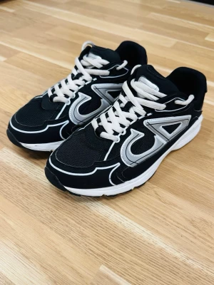Svarta sneakers med vita detaljer - Snygga svarta sneakers med vita och grå detaljer samt vita skosnören. Skorna har en sportig design med mesh och syntetmaterial, perfekt för en aktiv livsstil. Sulan är vit med svarta partier undertill och skon har en modern look.