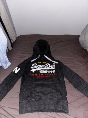 Mörkgrå Superdry hoodie med tryck - Snygg mörkgrå hoodie från Superdry med vit logga och text på bröstet samt färgglada detaljer. Huva med vita snören och tryck på ena ärmen. Perfekt för en avslappnad streetwear-look.