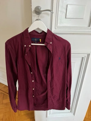 Vinröd skjorta från Ralph Lauren - Klassisk vinröd skjorta från Ralph Lauren med knappstängning och lång ärm. Skjortan har en broderad logga på bröstet och är tillverkad i bomull. Perfekt för en stilren och avslappnad look.