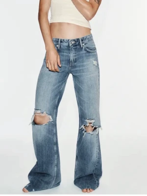 Blå slitna bootcut jeans - Säljer ett par blå bootcut jeans med stora slitningar över knäna och lätt tvättad look. Jeansen har hög midja, fem fickor och vida ben. Perfekta för en avslappnad och trendig stil.