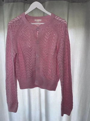 Rosa stickad kofta från Gina Tricot - Söt rosa stickad kofta från Gina Tricot med genombrutet mönster och stora knappar framtill. Koftan har långa ärmar, ribbade muddar och en mjuk, mysig känsla. Perfekt att slänga över en topp för en trendig look.