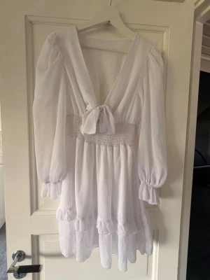 Vit kort omlottklänning med volang - Supersöt vit kort klänning med omlott och knyt framtill. Klänningen har långa puffärmar, smock i midjan och volangdetaljer längst ner. Perfekt för sommarens alla tillfällen och ger en romantisk vibe.