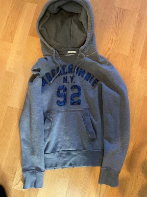 Grå hoodie Abercrombie N.Y. 92 - Snygg grå hoodie från Abercrombie med blå text och siffran 92 på bröstet. Klassisk känguruficka framtill och stor huva. Mjuk insida och ribbade muddar vid ärmar och nederkant. Perfekt för en avslappnad streetstyle-look.PRISET ÄR INTE HUGGET I STEN!