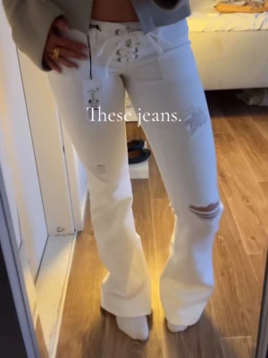 Vita bootcut jeans med snörning - Säljer ett par vita lågmidjade jeans med bootcut passform och cool snörning i midjan. Jeansen har slitna detaljer och hål på låret och knät, vilket ger en trendig look. Perfekta för dig som gillar att sticka ut och vill ha något unikt i garderoben.