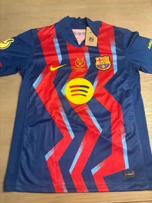 Barcelona matchtröja Nike blå/röd - Säljer en officiell FC Barcelona matchtröja i blått och rött med gula detaljer. Tröjan har klubbmärke, Nike-logga, sponsortryck och nummer 19 med namnet Roony på ryggen. Tillverkad i lätt och ventilerande material, perfekt för fotbollsträning eller match.