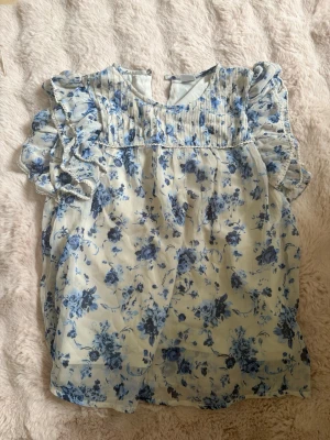 Blommig blus med volangärmar - Söt blus i tunn, ljus tyg med blått blommönster. Blusen har volangprydda korta ärmar och plisserad detalj vid bröstet. Rund halsringning och knapp i nacken. 