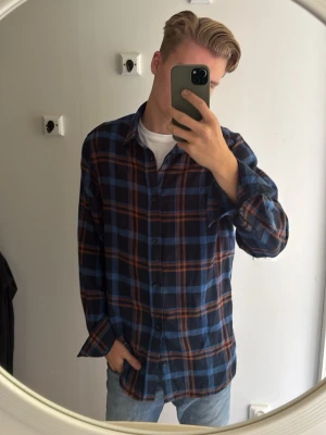 Flanell skjorta vintage - Säljer en oversized rutig skjorta i blått och orange med långa ärmar och knappar framtill. Skjortan har en avslappnad passform och är tillverkad i mjukt bomullstyg. Perfekt att bära över en t-shirt för en chill look. Flanell skjorta