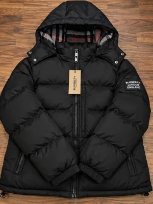 Svart pufferjacka från Burberry - Snygg svart pufferjacka från Burberry med huva och klassiskt rutigt foder i grått, rött och vitt. Jackan har dragkedja framtill, två fickor med dragkedja och Burberry-logga på ärmen. Perfekt för kalla vinterdagar och riktigt trendig look. Armar kan tas av så den blir väst.