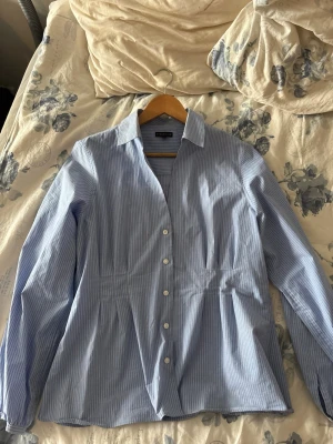 Blåvit randig skjorta  - Snygg blåvit randig skjorta med långa ärmar och klassisk krage. Skjortan har knappar framtill och lätt insvängd midja för en feminin siluett. Perfekt till jeans eller kjol för en fräsch look.