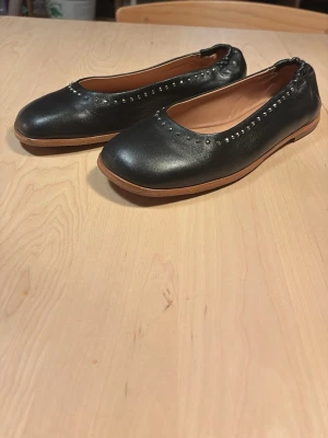 Svarta ballerinaskor med nitar - Säljer ett par svarta ballerinaskor i skinn med små silverfärgade nitar längs kanten. Skorna har rundad tå och ljusbrun sula i gummi. Perfekta för dig som gillar en enkel men ändå edgy stil.