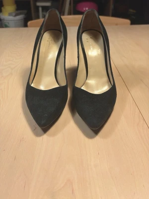 Svarta klassiska pumps i mocka - Snygga svarta pumps med spetsig tå och smal klack. Skorna har en elegant siluett och är tillverkade i mocka med beige innersula. Perfekta för dig som vill ha en stilren och tidlös look.