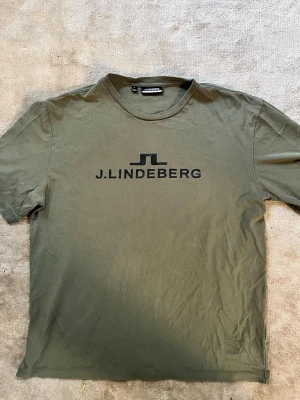 Olivgrön t-shirt från J.Lindeberg - Snygg olivgrön t-shirt från J.Lindeberg i storlek S med svart logga och text på bröstet.       Fraktar inom 24h🚚💨