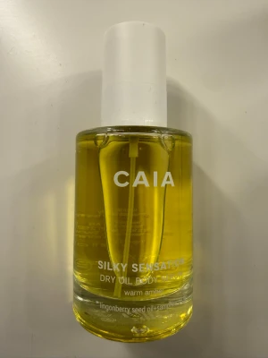 CAIA Silky Sensation Dry Oil Body -  kroppsolja från CAIA i en genomskinlig glasflaska med vit pump. Oljan har en varm gul färg och innehåller lingonfröolja och squalane. Doften är warm amber och flaskan rymmer 100 ml. Perfekt för att ge huden en silkeslen känsla.