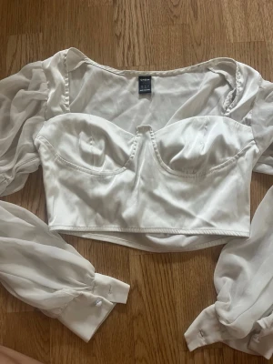 Vit korsettopp med puffärmar från SHEIN - Supersnygg vit korsettopp från SHEIN med transparenta puffärmar och markerade kupor. Toppen har croppad passform och knappar vid ärmsluten. Perfekt för dig som vill sticka ut med en trendig och feminin look.