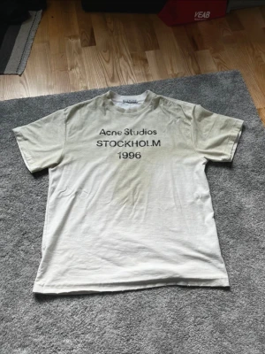  Acne Studios t-shirt - Säljer en beige t-shirt från Acne Studios med svart text 'Acne Studios STOCKHOLM 1996' på bröstet. Klassisk passform med rund hals och korta ärmar. Perfekt för dig som gillar minimalistisk och stilren design.
